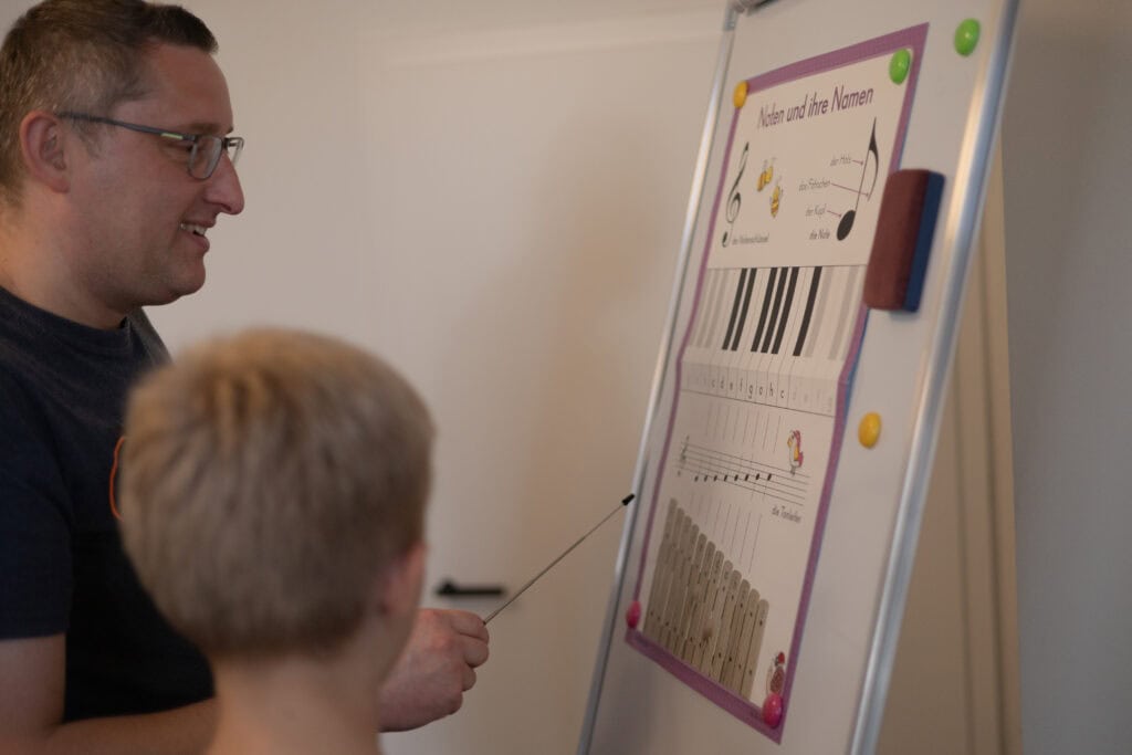 Kind zeigt am Flipchart auf ein Lernplakat mit Notenschlüssel, Notennamen und Klaviatur, die Lehrkraft schaut zu